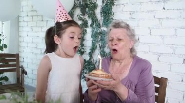 Ağır çekim. Mutlu küçük sevimli kız ve büyük anne 85. doğum gününü kutluyor, pastayı tutuyor ve birlikte mum üflüyor, beyaz tuğlalı arka plan. Tadını çıkar, saygı ve sıcak ilişki kavramı