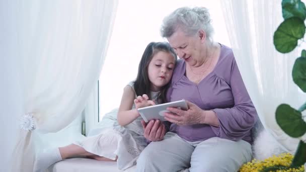 Bonito pequena menina pré-escolar ensinando a velha avó madura babá usando tablet digital. Família de duas gerações avô e neto explorando novas tecnologias pad dispositivo técnico em casa — Vídeo de Stock