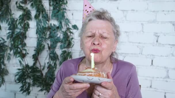 Lassú mozgás. Boldog mosolygó idősebb nagymama ünnepli 85. születésnapját, tartja torta és elfújja gyertya érzi hála, fehér tégla háttér. Élvezze, tisztelet és meleg kapcsolat. Ünnepi koncepció — Stock videók