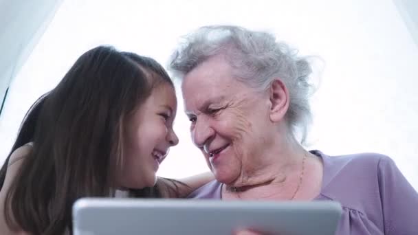 Маленькая дошкольница и старая взрослая бабуля-няня используют цифровые планшеты, улыбаются, обнимают друг друга. Два поколения семьи бабушки и дедушки и внуки исследуют новые технологии планшетных устройств техники на дому — стоковое видео