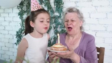 Mutlu küçük sevimli kız ve büyük anne 85. doğum gününü kutluyor, pastayı tutuyor ve birlikte mum üflüyor, beyaz tuğlalı arka plan. Tadını çıkar, saygı duy ve sıcak ilişki kur. Kutlama konsepti