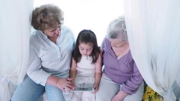 おばあちゃんやおばあちゃんのおばあちゃんと近くのデジタルタブレットを使用してかわいい小さな就学前の女の子。家庭で新しい技術パッドデバイス技術を探索する3世代の家族、コンピュータ中毒 — ストック動画