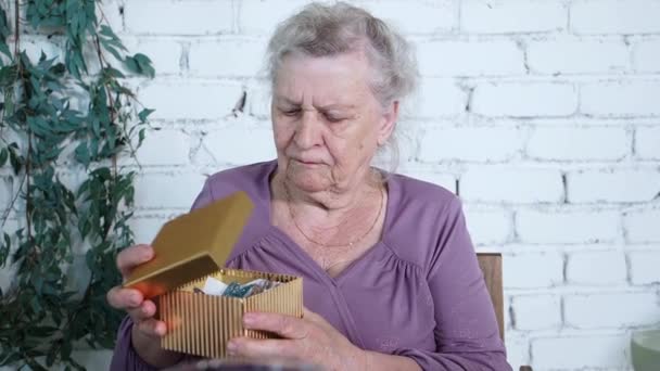 Šťastná starší žena drží dárek, otevírá krabici sedí u narozeninového stolu v obývacím pokoji. Usmívající se starší babička obdrží post zásilky on-line prodej objednávky spokojeni s rychlým dodáním koncept — Stock video