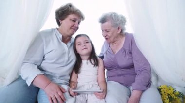Yaşlı büyük anne ve büyük anne dadı dijital tabletle küçük şirin bir anaokulu kızını öpüyor ve sarılıyor. Evde üç kuşak aile, kadın aile, sıcak bir ilişki ve saygı içinde.