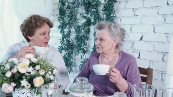 Zarif orta yaşlı bir kadın gülümseyerek masada yaşlı annesiyle çay içiyor, beyaz seramik bardakla sevgi ve şefkat dolu konuşuyorlar. Ailenin keyfi, saygısı ve sıcak ilişkisi — Stok video
