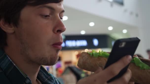 Mann isst Sandwich und checkt sein Handy in einem Restaurant in einem Einkaufszentrum. Nahaufnahme — Stockvideo