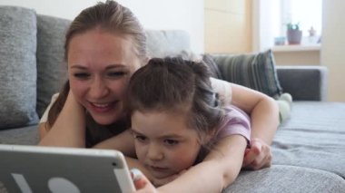 Mutlu aile annesi ve küçük kızı dijital tablet kullanarak çizgi film seyrediyor ve evdeki kanepede gülüyorlar. Sosyal uzaklık ve karantina karantinasında kişisel izolasyon sırasında.