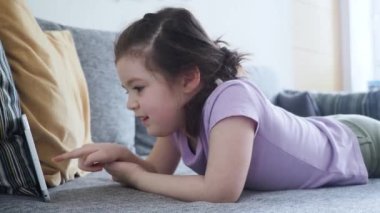 Kahverengi saçlı mutlu küçük kız evde dijital tablet kullanarak çizgi film izliyor. Sosyal uzaklık ve Coronavirus karantinası sırasında kanepede uzanıyor.
