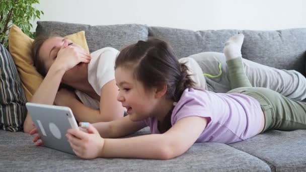 Cansada mãe boceja e adormece enquanto sua filha pequena usando tablet digital no sofá em casa. Menina aprende tecnologia durante a distância social e auto-isolamento em quarentena — Vídeo de Stock