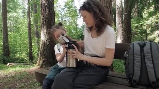 Путешественник мать и дочь открывают термос с горячим чаем сидя на скамейке во время лесного летнего похода. Счастливая семья наслаждается расслабляющим моментом во время отпуска. Время, проведенное вместе — стоковое видео