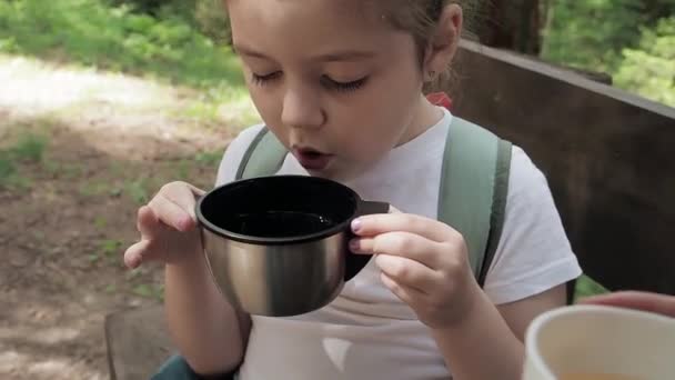 Kleine schattige reiziger meisje met lange zwarte wimpers en witte T-shirt blazen op hete thee in mok uit thermoskan tijdens bos zomerwandeling. Verfrissing en ontspannen moment tijdens familie vakantie reis — Stockvideo