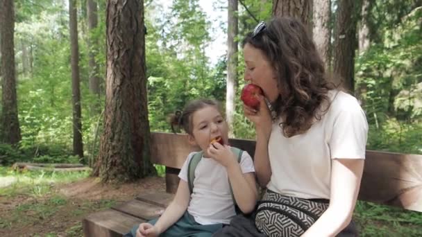 De cerca. Niño feliz hambriento con madre sentarse en el banco en el bosque y comer manzana roja para merendar en Camping Picnic al aire libre en el parque de senderos de la naturaleza. Viajar y pasar tiempo en familia juntos — Vídeos de Stock