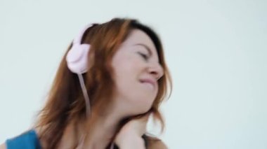 Pembe kulaklıklı genç, pozitif kızıl saçlı kadın müzik dinlerken gülümsüyor. Büyük kulaklıklara dokunur, beyaz stüdyo duvarında izole edilmiş şarkılardan ve danslardan zevk alır. Duygusal kadın portresi. Yakın plan.