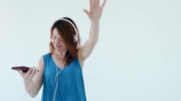 Retrato de una joven mujer pelirroja positiva con auriculares rosas y sonrisas de teléfono móvil mientras escucha música. Ella toca grandes auriculares, disfrutando de canciones y bailes aislados en la pared del estudio blanco — Vídeo de stock