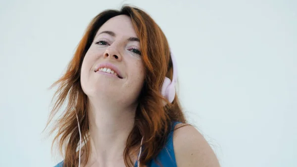Kapatın. Pembe kulaklıklı genç, pozitif kızıl saçlı kadın müzik dinlerken gülümsüyor. Beyaz stüdyo duvarında izole edilmiş şarkıları ve dansları seviyor. Duygusal kadın portresi