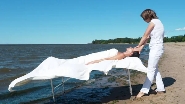 Genç bayan masaj terapisti yaz tatilinde kumsalda esmer kıza yüz masajı yapıyor. Masaj prosedürü. Genç bir bayan dışarıda masajdan hoşlanıyor. Sağlıklı yaşam tarzı ve vücut bakımı kavramı