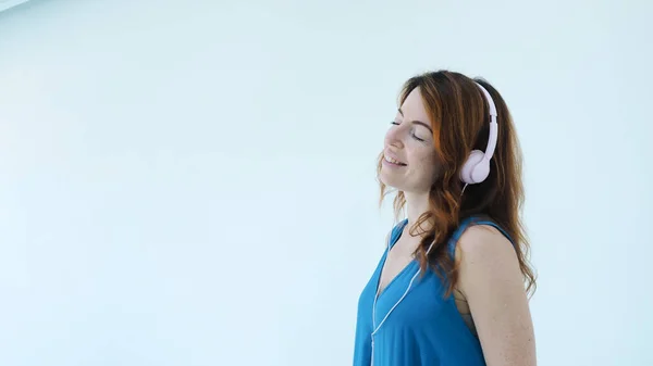 Pembe kulaklıklı genç, pozitif kızıl saçlı kadın müzik dinlerken gülümsüyor. Beyaz stüdyo duvarında izole edilmiş şarkıları ve dansları seviyor. Duygusal kadın portresi