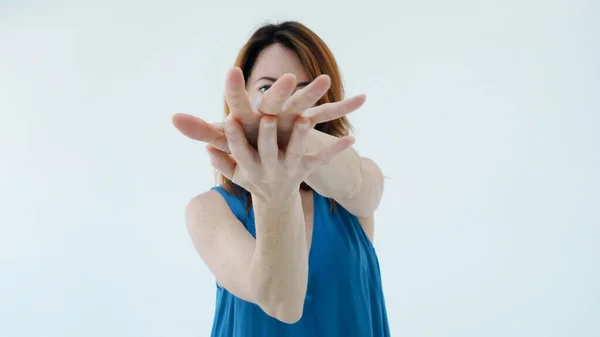 Kapatın. Mavi elbiseli güzel kızıl saçlı kadın yüzünü kapatıyor parmakları açık halde stüdyodaki beyaz duvarın arkasında tek başına duruyor. Güzellik konsepti