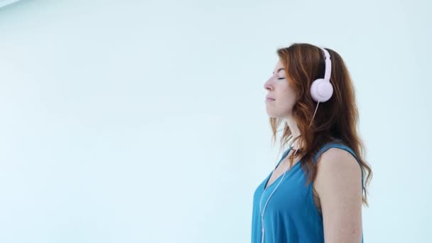 De cerca. Chica seria de pelo rojo con auriculares rosados y vestido azul escuchando música y bailando ligeramente con los ojos cerrados de lado. Disfrutando de canciones, meditación, tiempo a solas — Vídeos de Stock