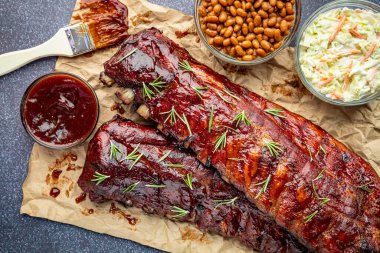 Kahverengi kağıda oturmuş iki raf kaburga, barbekü soslu fırça, bir kase sos, lahana salatası, fırında fasulye ve taze kekik otlarıyla süslenmiş.