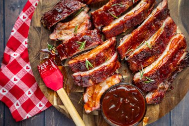 Barbekü kaburga parçaları ve yuvarlak ahşap bir kesme tahtasına yerleştirilmiş bir kase sos ve sos fırçası ve yanında kalpler olan kırmızı ve beyaz kareli peçete.