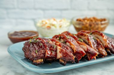 Açık mavi tabakta dilimlenmiş barbekü pirzolası. Yanında lahana salatası, arka planda fasulye ve bir kase sos.