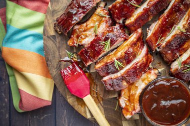 Barbekü kaburgaları dilimlenmiş ve yuvarlak ahşap bir kesme tahtasına yerleştirilmiş sos ve sos fırçası ve yanında parlak gökkuşağı renkli peçeteyle birlikte.