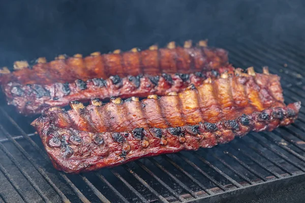 Barbekü soslu, ızgara tabaklarında pişen ve sigara içen kişinin görünür dumanıyla çevrili 2 sıra bbq kaburgasını kapatın.