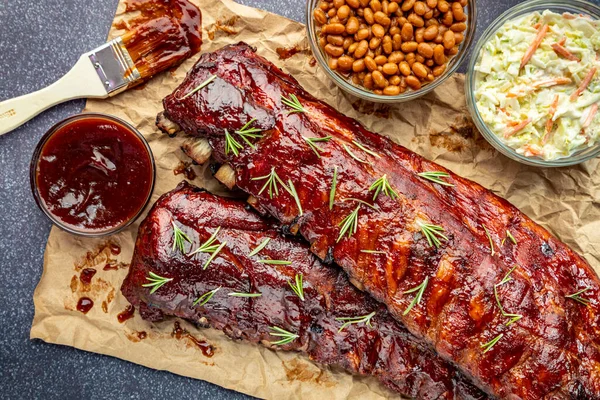 2 racks of ribs sitting on brown paper with barbeque sauce brush, sides of cole slaw, baked beans and garnished with fresh thyme herbs