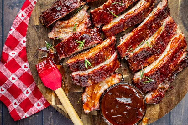 Barbekü kaburgaları dilimlenmiş ve yuvarlak ahşap bir kesme tahtasına yerleştirilmiş bir kase sos ve sos fırçası ve yanında kalpler olan kırmızı ve beyaz kareli peçete.