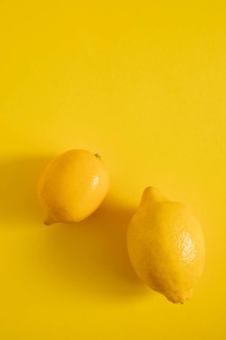 İki limon parlak sarı bir yüzeye oturur. Limonlar parlak bir ciltle yuvarlaktır. Arkaplan rengi meyvenin parlak ve taze görünümünü güçlendirir.