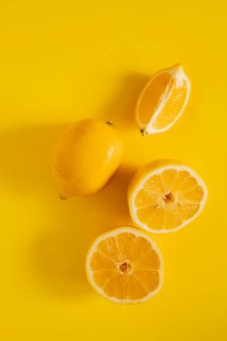 Parlak sarı bir yüzeyde bütün bir limon iki yarının yanında durur. Kesik meyve sulu içini gösteriyor. Düzenleme turunçgillerin tazeliğini temiz bir şekilde gösteriyor..
