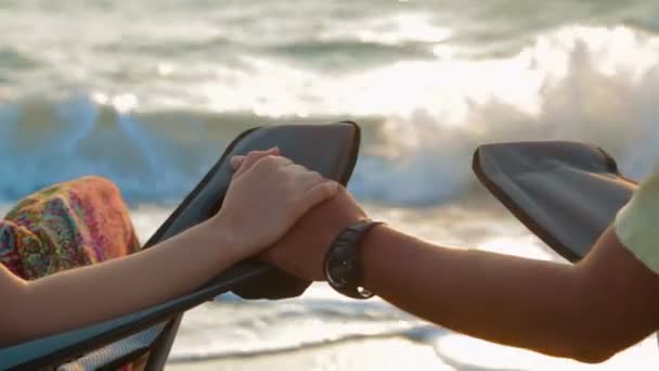 해변에서 야외 의자에 앉아있는 동안 손을 들고 사랑하는 커플 — 비디오