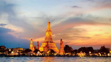 Wat Arun gün batımında altın ışıkta parlıyor, Tayland 'ın Bangkok şehrinde Chao Phraya nehrinin üzerinde dramatik bir akşam gökyüzü altında yansıyor..