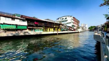 Geleneksel ahşap ve beton evler açık mavi gökyüzünün altında uzanan bir Bangkok kanalı, su yolundaki günlük şehir yaşamını yansıtıyor ve Tayland nehir kenarı mimarisi ve kültürünü gözler önüne seriyor..