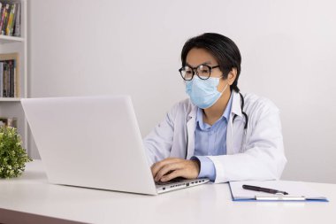 Laboratuvar önlüklü genç Asyalı doktor ya da gecelikli stetoskop, maske takıyor ve gözlük takıyor. Doktor Masasında Dizüstü bilgisayar kullanıyor.