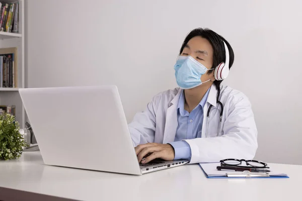 Relax Young Asian Doctor Man in Lab Coat or Gown with Stethoscope Wear Face Mask and Headphone Front Laptop Computer in Office