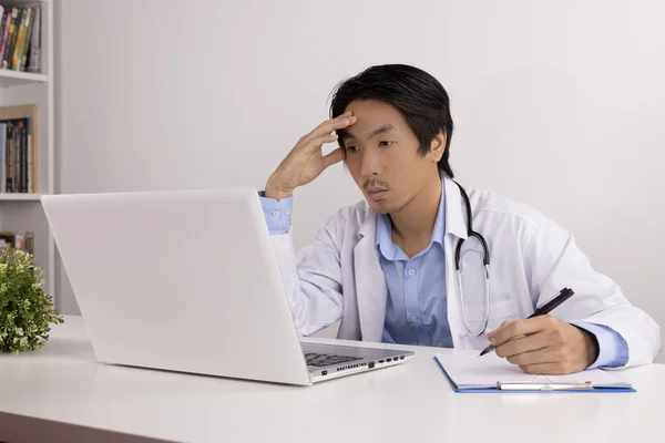 Serious Young Asian Doctor Man in Lab Coat or Gown with Stethoscope Writing Report and Using Laptop Computer on Doctor Table in Office