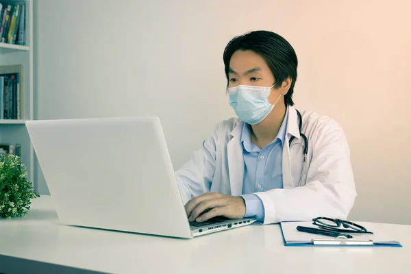 Young Asian Doctor Man in Lab Coat or Gown with Stethoscope Wear Face Mask Using Laptop Computer on Doctor Table in Office in Vintage Tone