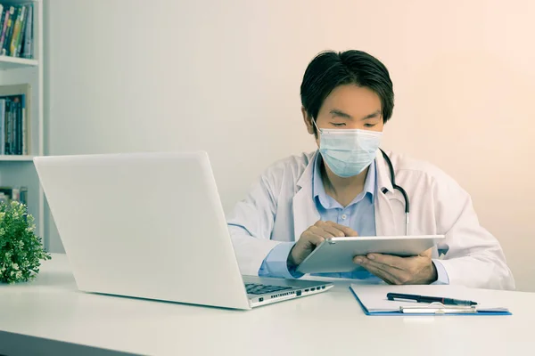 Young Asian Doctor Man in Lab Coat or Gown with Stethoscope Wear Face Mask Using Digital Tablet on Doctor Table in Office in Vintage Tone