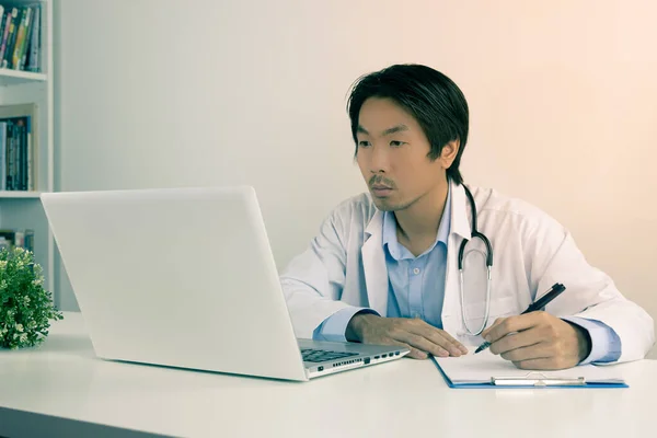 Young Asian Doctor Man in Lab Coat or Gown with Stethoscope Writing Experimental Results Report and Using Laptop Computer on Doctor Table in Office in Vintage Tone