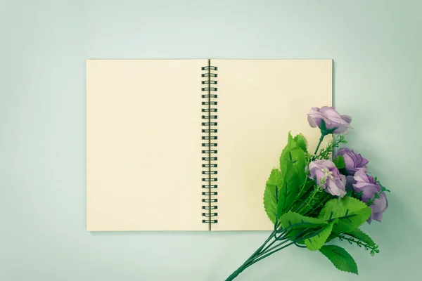 Top Table Spiral Notebook or Spring Notebook Mockup in Unlined Type on Center Frame and Purple Flowers at Bottom Right Corner on Blue Pastel Minimalist Background in Vintage Tone