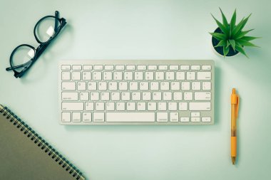 Mavi Pastel Minimalist Arkaplanda Beyaz Taşınabilir Bilgisayar Klavyesi veya Klavye Düğmesi ve Spiral Not Defteri ve Turuncu Kalem ve Ofis Bitkisi ve Gözlükleri