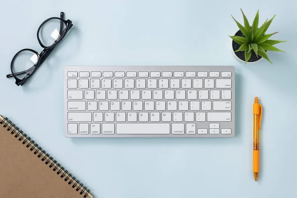 ホワイトポータブルコンピュータキーボードキーまたはキーボードボタンとスパイラルノートブックとオレンジペンとオフィスブルーパステルミニマリストの背景に植物と眼鏡 — ストック写真