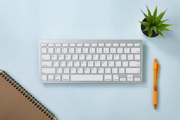 White Portable Computer Keyboard Keys or Keyboard Button and Spiral Notebook and Orange Pen and Office Plant on Blue Pastel Minimalist Background