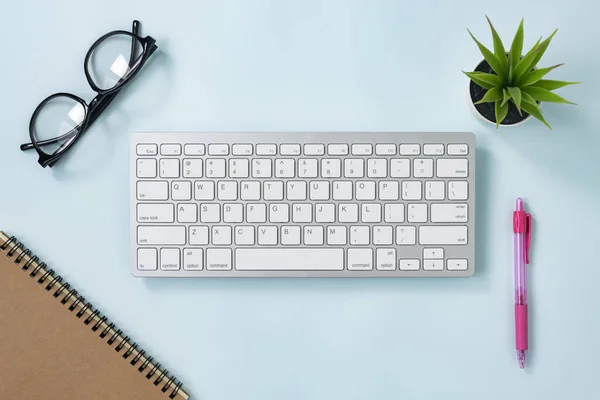 ホワイトポータブルコンピュータキーボードキーまたはキーボードボタンとスパイラルノートブックとピンクペンとオフィスブルーパステルミニマリストの背景に植物と眼鏡 — ストック写真