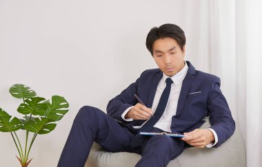 Asian Financial Advisor Check Financial Statement in Clipboard on Bean Bag in Home Office. Businessman Work at Home for Social Distance or Home Quarantine in Covid 19 or Coronavirus Situation