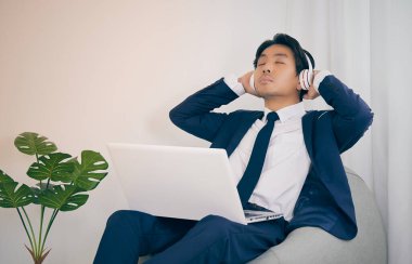 Asyalı iş adamı müzik dinlemek için dizüstü bilgisayar kullanarak kulaklık takıyor. Covid 19 'da Rahat Çalışma Saati ya da Ev Karantinası ya da Klasik Ton' da Coronavirüs Durumu