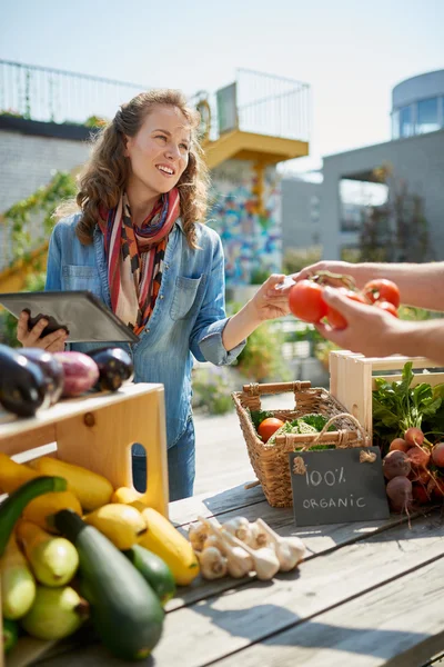 Friendly woman tending an organic vegetable stall at a farmers market and selling fresh vegetables from the rooftop garden Stock Picture