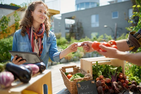 Vänlig kvinna tenderar en ekologisk vegetabiliska stall på en farmers market och säljer färska grönsaker från takträdgården Royaltyfria Stockfoton